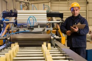 Vooraanzicht van medewerker in een fabriek met gele helm op, blauwe outfit en een klembord in zijn hand, naast een grote machine.